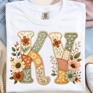 Puede incluir: Camiseta blanca con las letras "KV" en un diseño floral de patchwork. Las letras están adornadas con flores, hojas y bayas en tonos naranja, amarillo, verde y rosa. La camiseta tiene cuello redondo y una etiqueta "Comfort Colors".
