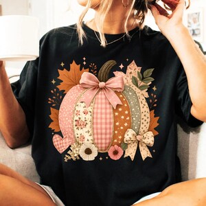 May include: Black t-shirt featuring a colorful pumpkin design. The pumpkin is composed of various patterns, including floral, gingham, and star prints, with a pink bow and autumn leaves. The design includes a heart and flowers.