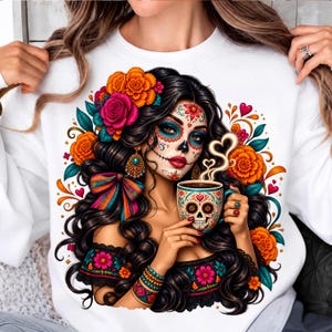 May include: A white sweatshirt featuring a graphic print of a person with long dark hair, wearing floral hair accessories and sugar skull makeup. The person holds a mug with a skull design and steam rising in heart shapes, surrounded by orange flowers.