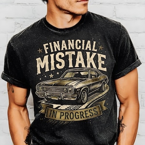 Könnte beinhalten: Schwarzes T-Shirt mit einem Used-Look-Grafikdesign. Das Design zeigt die Worte "FINANCIAL MISTAKE" über einer Vintage-Auto-Illustration, mit "IN PROGRESS" auf einem Banner darunter. Das T-Shirt hat einen klassischen Rundhalsausschnitt und kurze Ärmel.