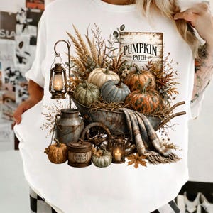 May include: A white t-shirt featuring a graphic print of a rustic wheelbarrow filled with various pumpkins, dried wheat, and a plaid blanket. The design includes a metal lantern, a milk can, a container labeled Farm Fresh, and a sign reading PUMPKIN PATCH.