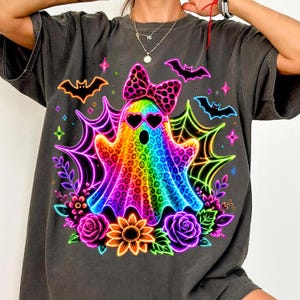 May include: A charcoal grey t-shirt featuring a vibrant, rainbow-coloured ghost graphic with a leopard print pattern. The ghost wears a matching leopard print bow and heart-shaped sunglasses, surrounded by neon spiderwebs, bats, and flowers.
