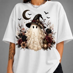 K&ouml;nnte beinhalten: Ein wei&szlig;es T-Shirt mit einer Grafik eines Geistes, der einen schwarzen Hexenhut tr&auml;gt, umgeben von dunkelroten und schwarzen Rosen, einer leuchtenden Laterne, einer Mondsichel und kleinen Flederm&auml;usen auf einem spitzenartigen Stoffmuster.