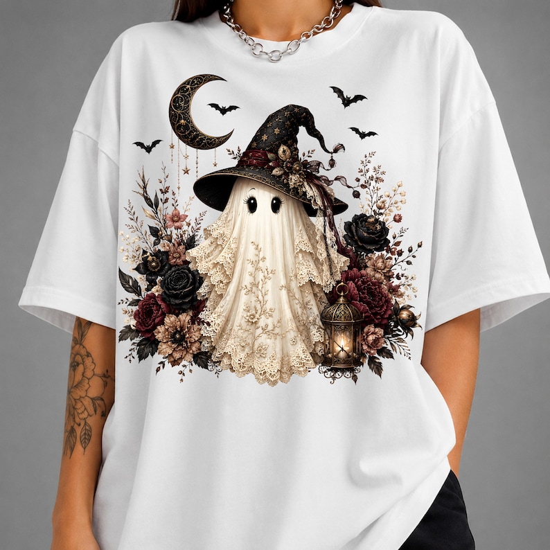 K&ouml;nnte beinhalten: Ein wei&szlig;es T-Shirt mit einer Grafik eines Geistes, der einen schwarzen Hexenhut tr&auml;gt, umgeben von dunkelroten und schwarzen Rosen, einer leuchtenden Laterne, einer Mondsichel und kleinen Flederm&auml;usen auf einem spitzenartigen Stoffmuster.