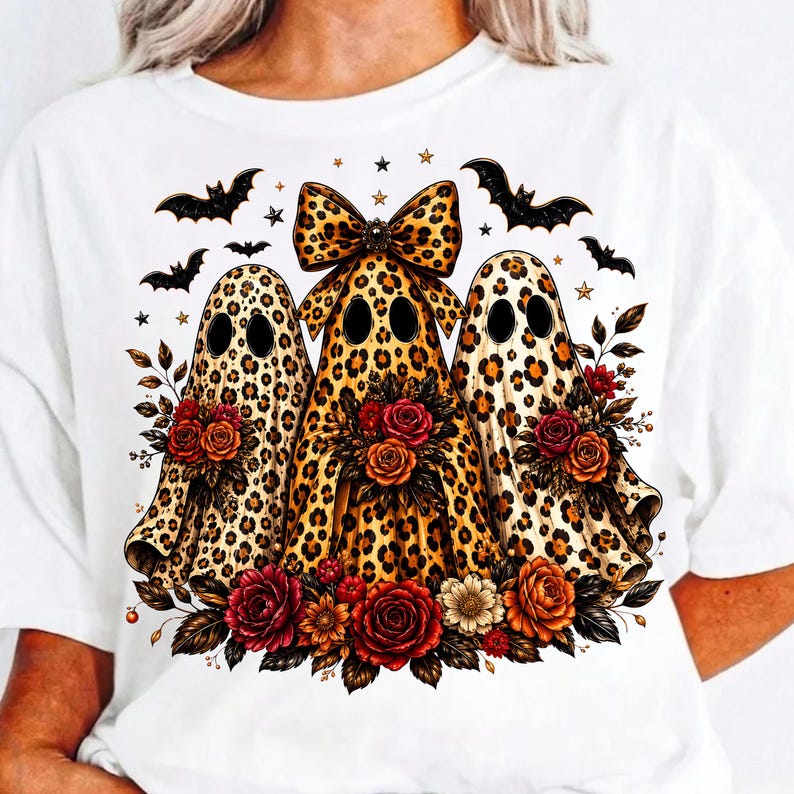 May include: A white t-shirt featuring a graphic of three ghosts with a brown and black leopard print pattern. The center ghost wears a large leopard print bow. Each ghost is surrounded by red, orange, and white flowers, with black bats and small stars.