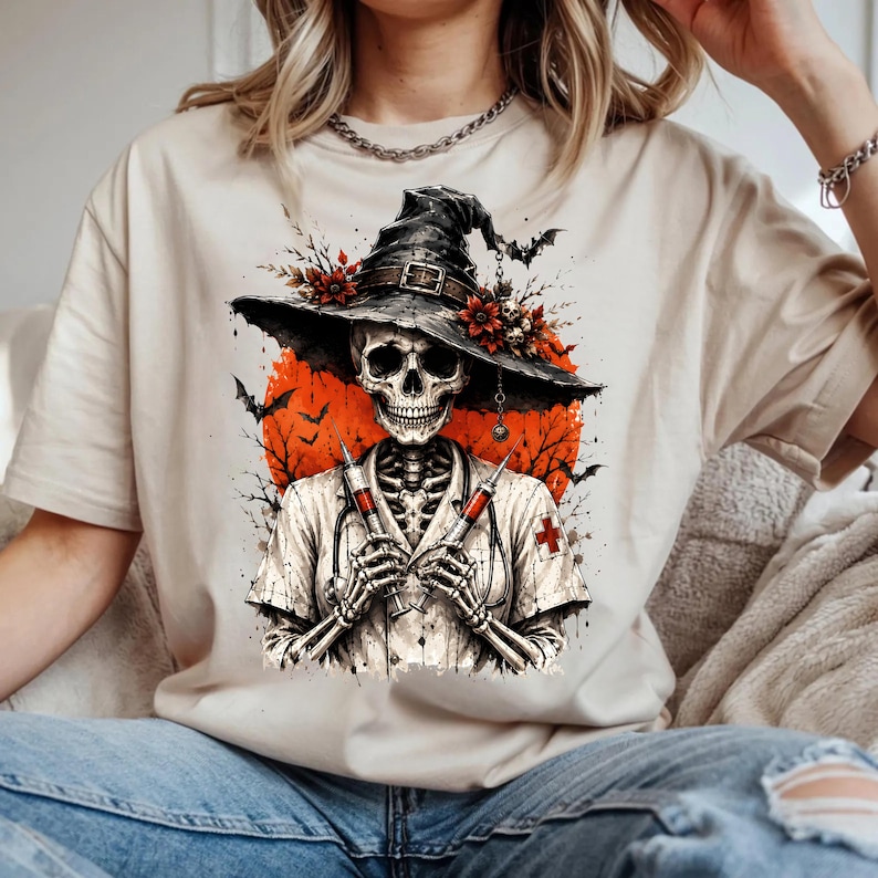 May include: A beige t-shirt featuring a graphic of a skeleton dressed as a nurse, wearing a wide-brimmed black hat adorned with red flowers and small skulls. The skeleton holds two syringes, set against a large orange moon with black bat silhouettes.