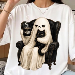 May include: A white t-shirt featuring a graphic of a ghost sitting in a black armchair. The ghost holds a black mug with the text MEH and cradles a black cat. The ghost has a sad facial expression, and steam rises from the mug in the illustration.