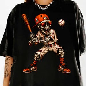 May include: A black t-shirt featuring a graphic of a skeleton baseball player wearing an orange cap with a skull emblem, sunglasses, and a blood-splattered uniform. The skeleton holds a spiked wooden bat, ready to hit a baseball floating nearby.