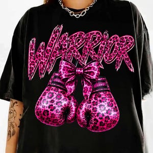 May include: A black t-shirt featuring a graphic print of two pink and black leopard-print boxing gloves tied together with a matching bow. Above the gloves, the word WARRIOR is printed in a large, stylized pink and black leopard-print font.
