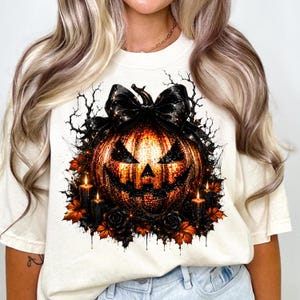 May include: A cream-colored t-shirt featuring a graphic of a glowing jack-o'-lantern topped with a large black bow. The pumpkin is surrounded by black candles, dark roses, orange autumn leaves, and bare tree branches on a white background.