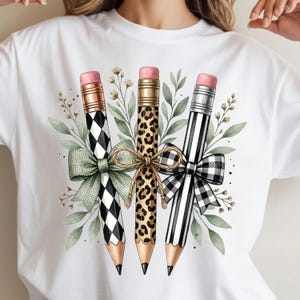 May include: A white t-shirt featuring a graphic of three pencils with pink erasers. The pencils have black and white diamond, leopard print, and black and white stripe patterns. They are decorated with green and plaid bows and green leaves.