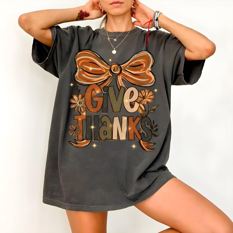 May include: Dark gray t-shirt with a large bow graphic in shades of brown and orange. The words "GIVE THANKS" are printed in a fall-themed font, surrounded by floral accents. A casual, oversized fit.