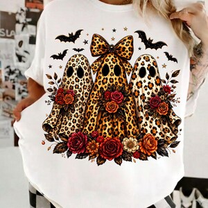 May include: A white t-shirt featuring a graphic of three ghost figures with a leopard print pattern. The central ghost wears a large leopard print bow. The ghosts are surrounded by black bats, small stars, and clusters of red and orange roses.