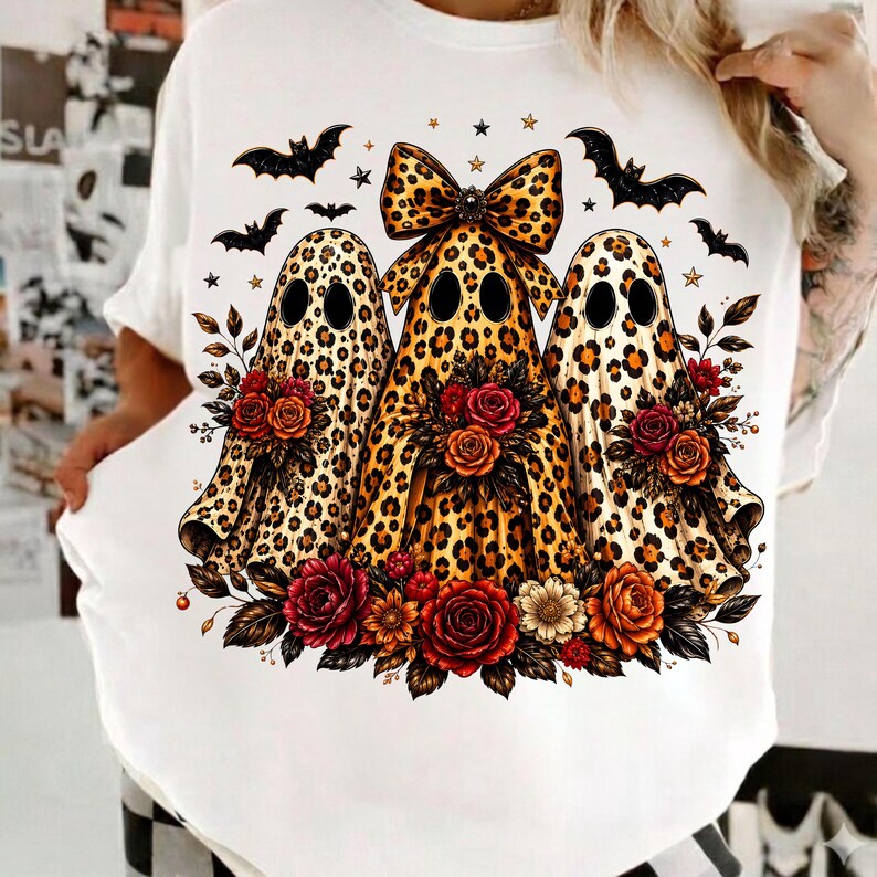 May include: A white t-shirt featuring a graphic of three ghost figures with a leopard print pattern. The central ghost wears a large leopard print bow. The ghosts are surrounded by black bats, small stars, and clusters of red and orange roses.