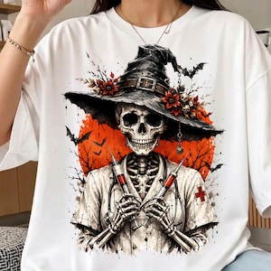 May include: A white t-shirt featuring a graphic print of a skeleton dressed as a nurse and wearing a witch hat. The skeleton holds two syringes, with a stethoscope around its neck, set against a large orange moon, bats, and bare tree branches.