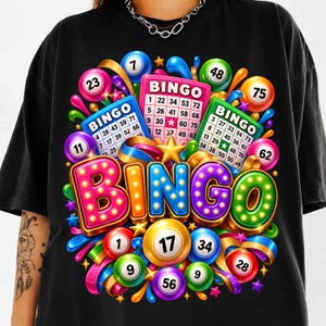 May include: A black t-shirt featuring a vibrant graphic print of the word BINGO in large, colourful, illuminated-style letters. The design includes multiple numbered bingo balls in various colours and three bingo cards with printed numbers.