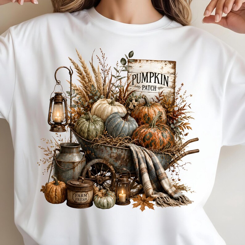 Może przedstawiać: Biały t-shirt z grafiką metalowej taczki wypełnionej pomarańczowymi, zielonymi i niebieskimi dyniami, suszoną pszenicą, latarnią oraz znakiem z napisem PUMPKIN PATCH. Obok znajduje się metalowa bańka na mleko i słoik z napisem FARM FRESH.