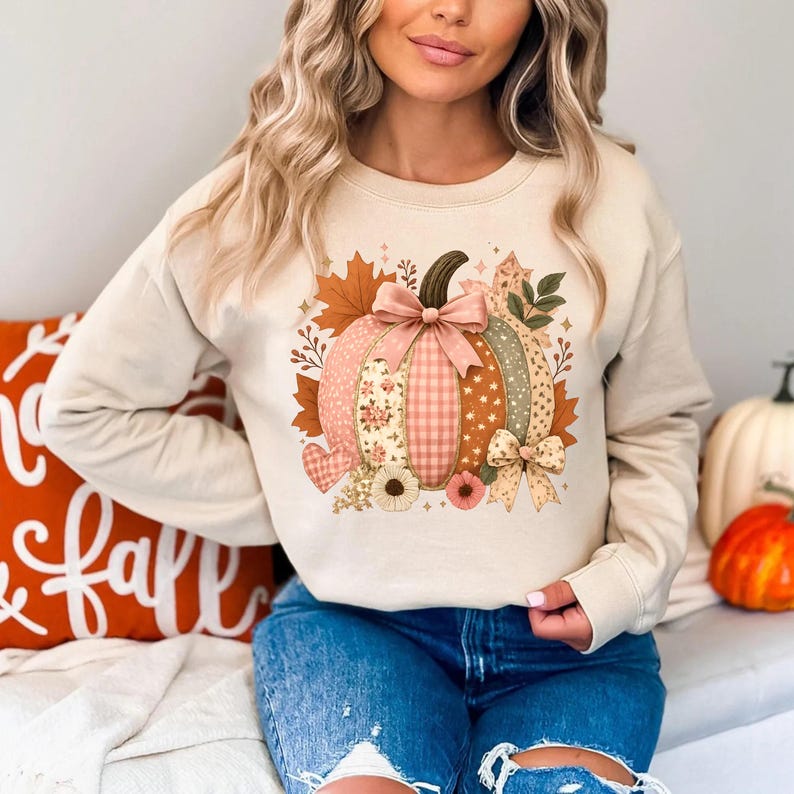May include: A beige sweatshirt featuring a colorful pumpkin design. The pumpkin is composed of various patterns, including floral, gingham, and polka dots, with a pink bow. Autumn leaves and floral accents surround the pumpkin, creating a fall theme.