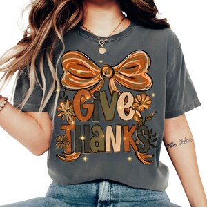 May include: A gray t-shirt with a large graphic that says "Give Thanks" in a fall-themed design. The design includes a bow, flowers, and leaves in shades of orange, brown, and green. The shirt is a comfortable, casual style.