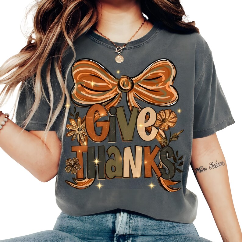 May include: A gray t-shirt with a large graphic that says "Give Thanks" in a fall-themed design. The design includes a bow, flowers, and leaves in shades of orange, brown, and green. The shirt is a comfortable, casual style.
