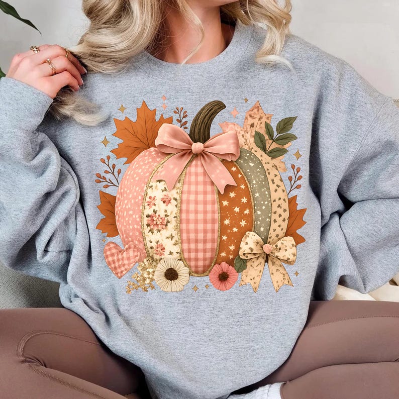 May include: A gray sweatshirt featuring a patchwork pumpkin design. The pumpkin has sections of floral, gingham, and star patterns, with a brown stem and a pink bow. Autumn leaves and flowers surround the pumpkin, creating a fall theme.
