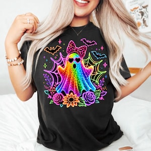 May include: A black t-shirt featuring a vibrant, neon-coloured graphic of a ghost with a leopard print pattern, heart-shaped sunglasses, and a polka-dot bow. The design includes glowing spiderwebs, bats, roses, and a sunflower in bright colours.