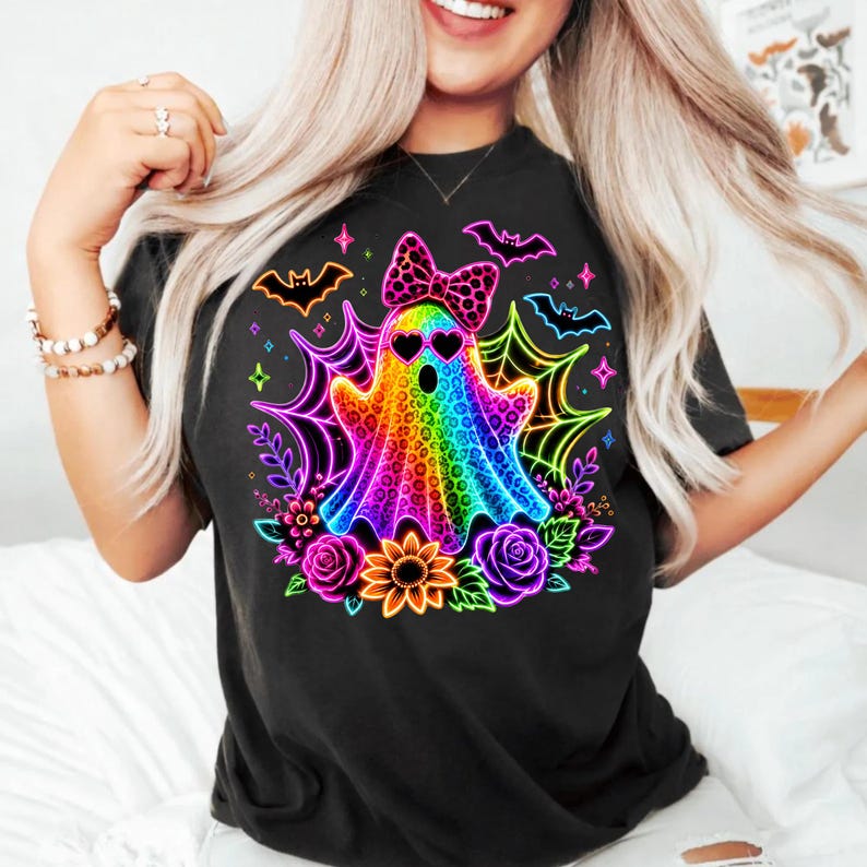 May include: A black t-shirt featuring a vibrant, neon-coloured graphic of a ghost with a leopard print pattern, heart-shaped sunglasses, and a polka-dot bow. The design includes glowing spiderwebs, bats, roses, and a sunflower in bright colours.