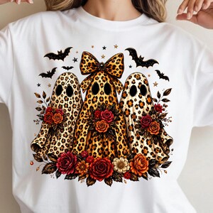 May include: A white t-shirt featuring a graphic of three ghosts with a brown and black leopard print pattern. The center ghost wears a large leopard print bow. The ghosts are surrounded by red and orange roses, black bats, and small stars.