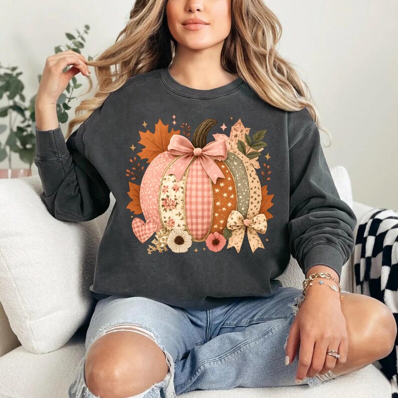 May include: Dark gray sweatshirt featuring a patchwork pumpkin design. The pumpkin has sections of floral, gingham, and dotted patterns, with a pink bow and autumn leaves. The design includes a heart and sunflowers, perfect for fall.