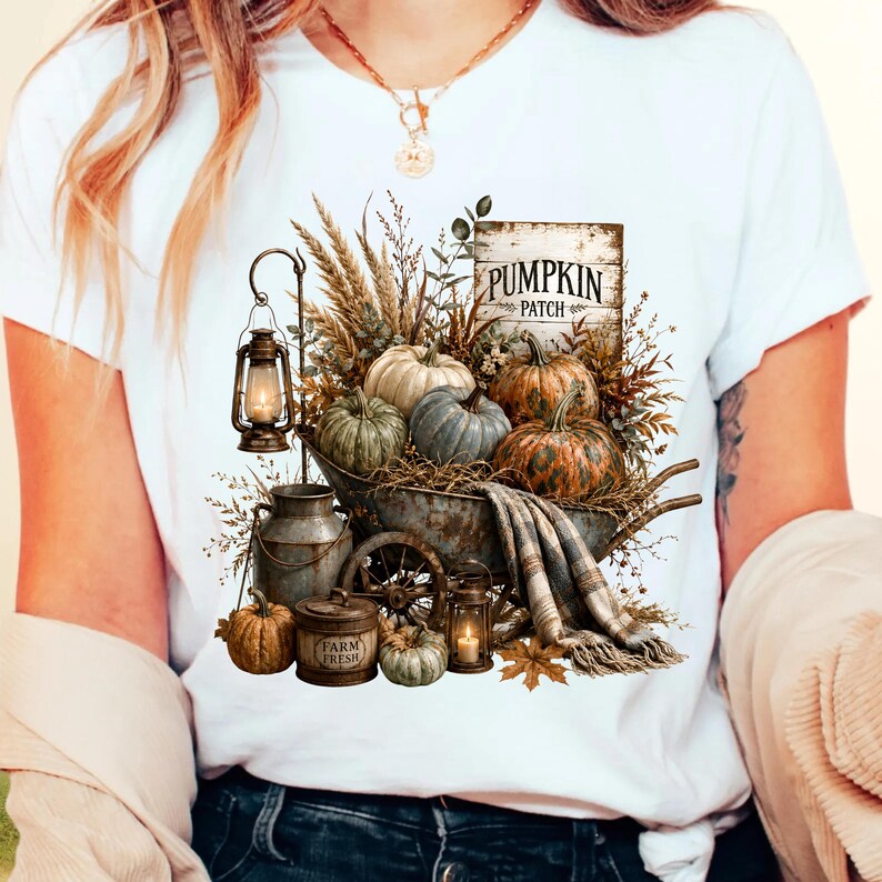 Może przedstawiać: Biały t-shirt z nadrukiem graficznym przedstawiającym rustykalną taczkę wypełnioną dyniami, suszoną pszenicą i znakiem z napisem PUMPKIN PATCH. Wz&oacute;r zawiera metalową latarnię, bańkę na mleko z napisem FARM FRESH oraz koc w kratę przewieszony przez bok.