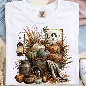 Może przedstawiać: Biały t-shirt z nadrukiem graficznym przedstawiającym rustykalną taczkę wypełnioną dyniami, suszoną pszenicą i latarnią. Drewniany znak ma napis PUMPKIN PATCH, a metalowy pojemnik jest oznaczony napisem FARM FRESH. Koc w kratę zwisa z boku.