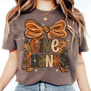 May include: A gray t-shirt featuring a large bow design in shades of brown and orange, with the words "GIVE THANKS" in a decorative font. The design includes floral accents and sparkling stars, perfect for the Thanksgiving season.