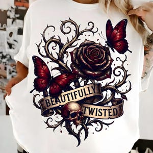 May include: White sweatshirt with a gothic-style graphic. The design features a dark red rose, butterflies, and a skull, with the text "BEAUTIFULLY TWISTED" on a banner. The artwork has a floral and dark theme.