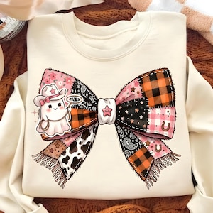 May include: Cream-colored sweatshirt featuring a large, patchwork bow design with a ghost wearing a cowboy hat. The bow incorporates pink, orange, black, and white patterns, including plaid, paisley, and cow print. A horseshoe and star accents are also visible.