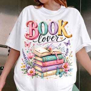 May include: A white t-shirt featuring a graphic print with the text "BOOK lover" in colorful, dimensional lettering. Below the text is a stack of four books surrounded by various flowers, lavender sprigs, and two butterflies in shades of pink, blue, and yellow.