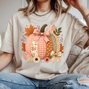 May include: Beige t-shirt featuring a patchwork pumpkin design with floral, gingham, and star patterns. The pumpkin has a brown stem and is adorned with pink bows and autumn leaves. A heart and flowers are also included in the design.