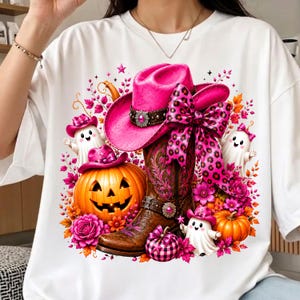 May include: A white t-shirt featuring a graphic print of a bright pink cowboy hat with a leopard print bow, a brown leather cowboy boot, orange pumpkins, and small white ghosts, all surrounded by pink and orange flowers and stars.