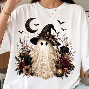 K&ouml;nnte beinhalten: Ein wei&szlig;es T-Shirt mit einer Grafik eines Geistes, der einen mit Blumen verzierten schwarzen Hexenhut tr&auml;gt. Der Geist ist umgeben von dunklen Rosen, einem h&auml;ngenden Halbmond, fliegenden Flederm&auml;usen und einer leuchtenden Laterne.
