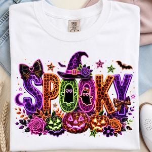 Op de afbeelding: Een wit t-shirt met het woord SPOOKY in grote, kleurrijke letters met luipaardprint. Het ontwerp bevat een paarse heksenhoed, twee oplichtende spooksilhouetten, drie pompoenen, zwarte strikken, vleermuizen, sterren en paarse en oranje rozen.
