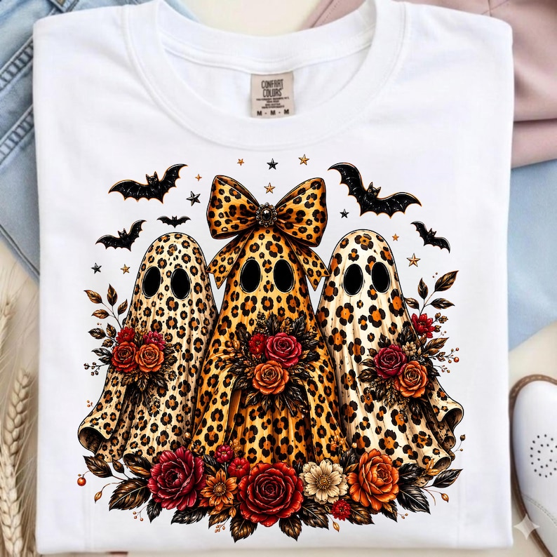 May include: A white t-shirt featuring a graphic of three ghosts with a brown and black leopard print pattern. The center ghost wears a large leopard print bow. Each ghost holds a bouquet of red, orange, and yellow roses. Black bats and small stars surround the ghosts.