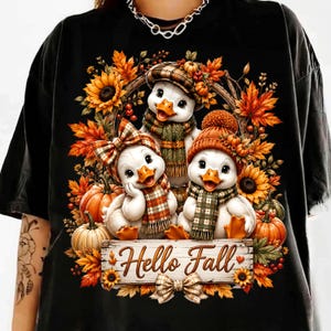 May include: A black t-shirt featuring a graphic of three white ducklings wearing scarves and hats, surrounded by autumn leaves, sunflowers, and pumpkins. A wooden sign at the bottom displays the text "Hello Fall" with a small bow underneath.