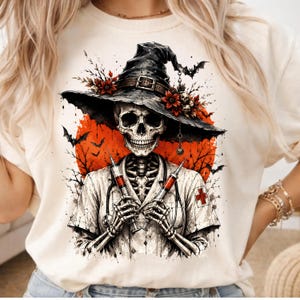May include: A cream-colored t-shirt featuring a graphic of a skeleton dressed as a nurse, wearing a witch hat adorned with red flowers and a skull. The skeleton holds two syringes, set against a large orange moon with silhouettes of bats and trees.