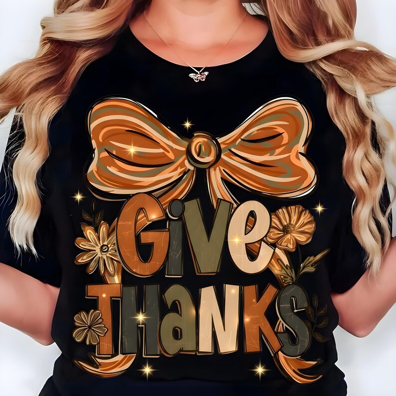 May include: Black t-shirt with a large bow and the words "GIVE THANKS" in a fall-themed design. The design includes floral elements and a color palette of orange, brown, and green. The shirt is a festive apparel item.