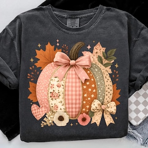 May include: Dark gray sweatshirt featuring a colorful pumpkin design. The pumpkin is segmented with floral, gingham, and star patterns, adorned with bows and surrounded by fall leaves and flowers. A heart and stars complete the autumn theme.