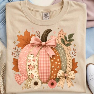 May include: Beige Comfort Colors t-shirt featuring a colorful pumpkin design. The pumpkin has sections of pink floral, gingham, and star patterns, with a brown stem and pink bows. Autumn leaves and flowers surround the pumpkin, creating a fall theme.