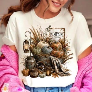 Może przedstawiać: Biały t-shirt z grafiką przedstawiającą rustykalną taczkę wypełnioną dyniami, suszoną pszenicą i latarnią. Znak głosi PUMPKIN PATCH, a mały pojemnik jest oznaczony jako FARM FRESH. Na boku taczki przewieszony jest koc w kratę o stonowanych barwach.