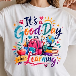 May include: A white t-shirt featuring a colorful graphic with the text It's a Good Day for Learning. The design includes a pink backpack, a blue pencil holder with pencils, a red apple, a globe on a stack of books, and various hearts and stars.
