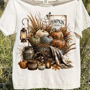 Może przedstawiać: Kremowy t-shirt z nadrukiem graficznym przedstawiającym rustykalną taczkę wypełnioną dyniami, suszoną pszenicą i kocem w kratę. Wz&oacute;r zawiera dwie latarnie, metalową bańkę na mleko, pojemnik z napisem FARM FRESH oraz znak PUMPKIN PATCH.