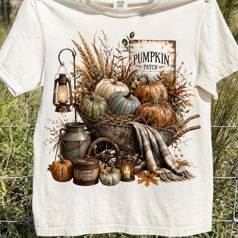 Może przedstawiać: Kremowy t-shirt z nadrukiem graficznym przedstawiającym rustykalną taczkę wypełnioną dyniami, suszoną pszenicą i kocem w kratę. Wz&oacute;r zawiera dwie latarnie, metalową bańkę na mleko, pojemnik z napisem FARM FRESH oraz znak PUMPKIN PATCH.