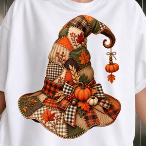 May include: A white t-shirt featuring a graphic of a patchwork witch hat. The hat is composed of various fabric patterns including plaid, gingham, and burlap in orange, green, and brown. It is decorated with pumpkins, leaves, and berries.